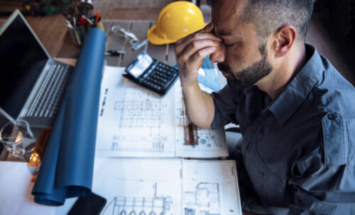 Overworked stressed male contractor having headache while sitting at desk full of drawings